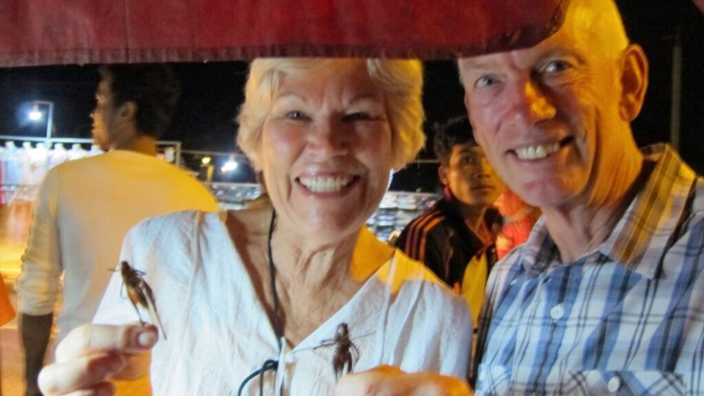 Older couple laughing while holding fried cricket snacks at a Siem Reap night market during the After Dark Food Tour