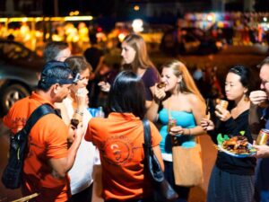 Clients et guides Adventures Cambodia en chemises orange partageant des brochettes de street-food grillées dans un marché de nuit à Siem Reap lors du After Dark Food Tour