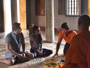 Local guide in an orange shirt explaining a pagoda ritual to two seated guests as a Buddhist monk looks on inside a Siem Reap temple