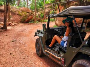 Akim Ly in a green hat behind the wheel of a vintage military jeep on a red dirt track between sandstone boulders
