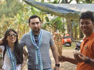 Local guide in an orange shirt offering a bunch of skewered crickets and tarantulas to a couple wearing krama scarves during a Siem Reap countryside tour