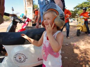 Small child smiling while trying on an oversized orange Vespa helmet beside a row of Vespas on a Siem Reap street