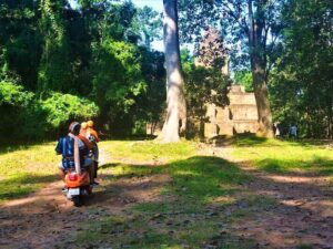 Twee gasten op een oranje Vespa die pauzeren op een zonnige open plek in het bos voor een kleine Angkor-tempelruïne