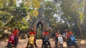 Group of guests in orange vests on colourful Vespas posing in front of a carved stone gateway at Angkor