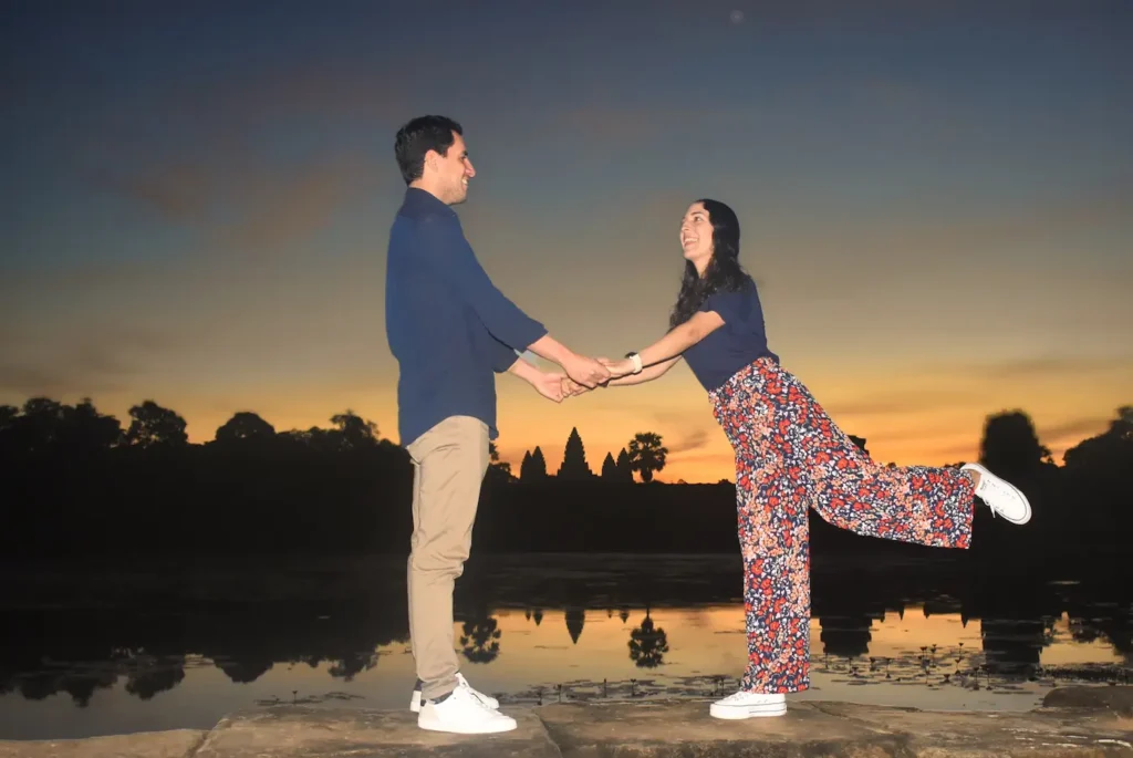Akim Ly and her husband holding hands and laughing on the moat wall at Angkor Wat at sunrise, temple silhouette reflected in the water
