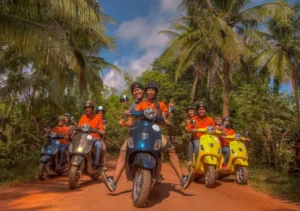 Group of tourists in orange shirts riding colorful Vespas on a red dirt road through palm trees near Siem Reap