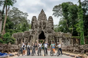 Group of cyclists with raised arms posing on bikes in front of the stone towers of Angkor Thom's gate entrance