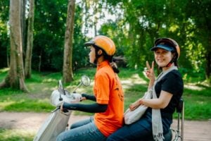 Two women on a Vespa scooter wearing helmets in a tree-lined road near Siem Reap