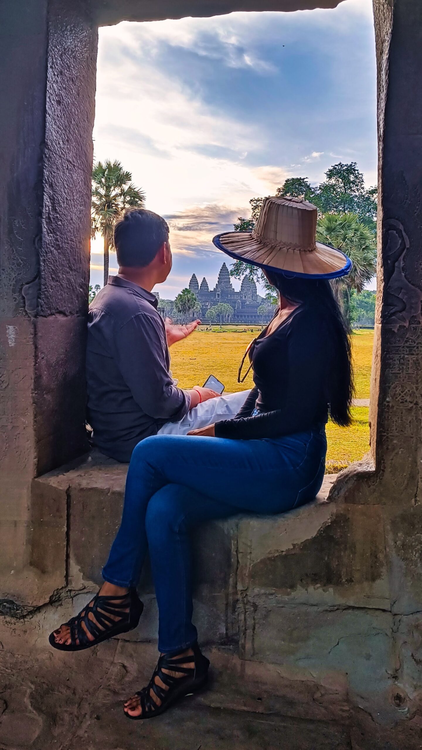 Couple sitting in ancient stone window frame watching Angkor Wat towers at sunrise
