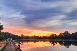 Visitors stand on a pathway at sunrise beside Angkor Wat's moat, reflected in calm water under orange and pink sky