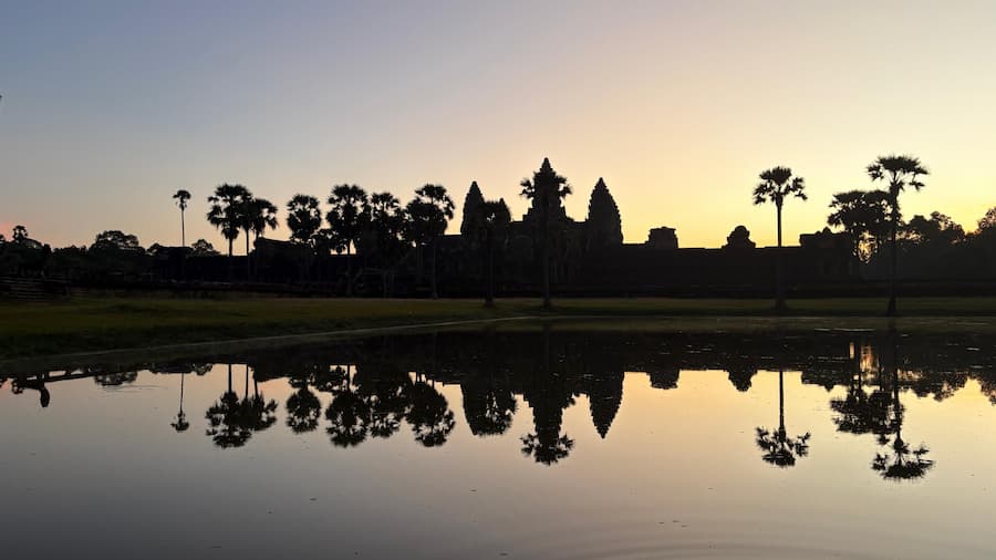 Angkor Wat temple silhouetted at sunrise with palm trees reflected in the southern moat