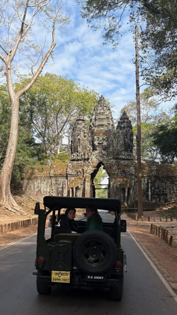 A vintage Willys jeep on a tree-lined road to an Angkor gate