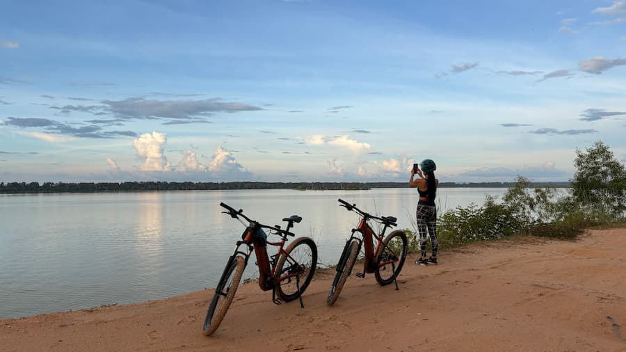 Two pedal-assist bikes on the red sand shore of the West Baray while a rider photographs the water