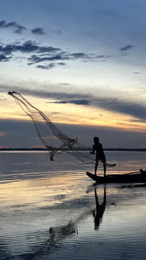 A Cambodian fisherman casts a net from a wooden boat at sunset on the West Baray