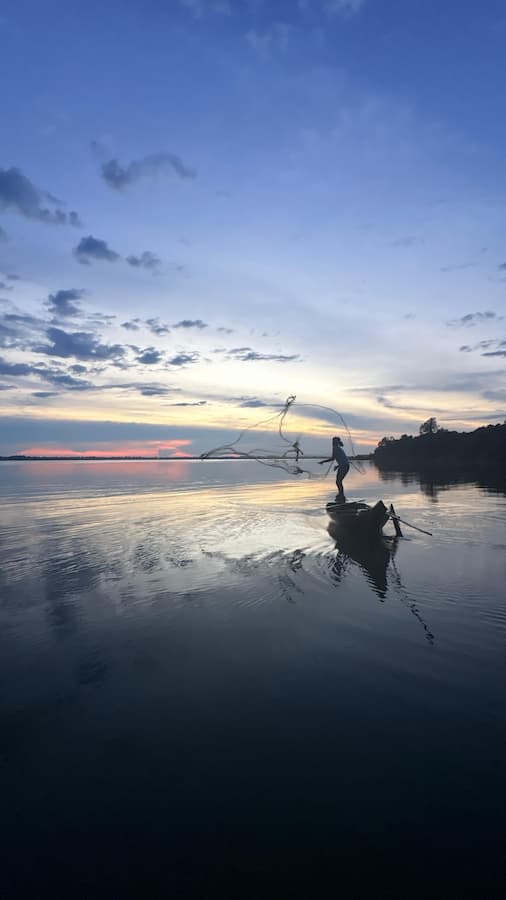 A Cambodian fisherman throws a net from a wooden boat at dusk on the West Baray