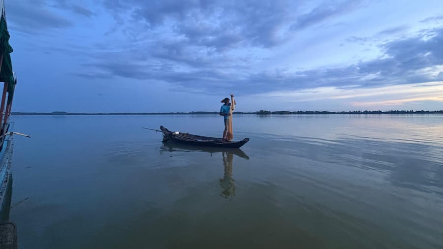 Visser die in een houten boot staat op het rustige water van West Baray bij schemering onder een blauwe lucht