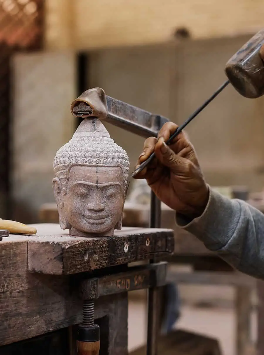 An artisan carving a sandstone Buddha head with a chisel and mallet at Artisans Angkor, Siem Reap
