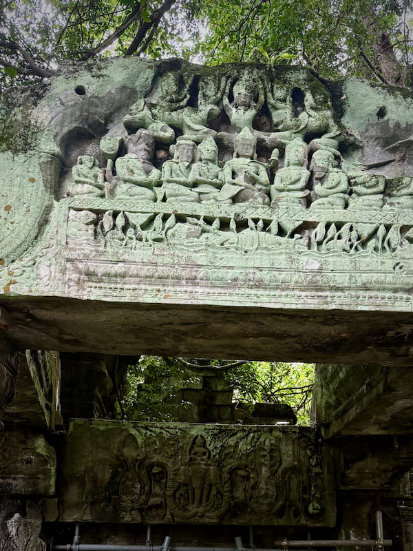 A carved pediment of seated figures above a decorated lintel at Beng Mealea, green lichen across the stone