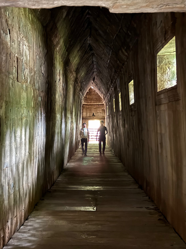 Visitors silhouetted in the corbelled stone gallery at Beng Mealea, walking the wooden floor toward the light