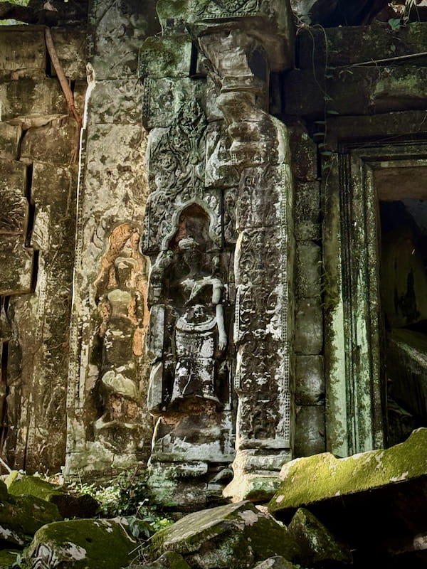 A carved devata standing in her niche on a pilaster at Beng Mealea, moss and lichen across the sandstone