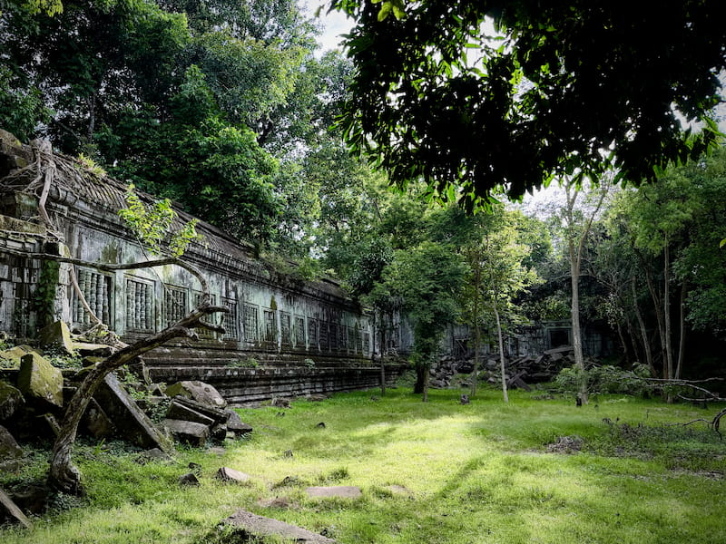 A long gallery wall with baluster windows beside sunlit green grass at Beng Mealea in the rainy season