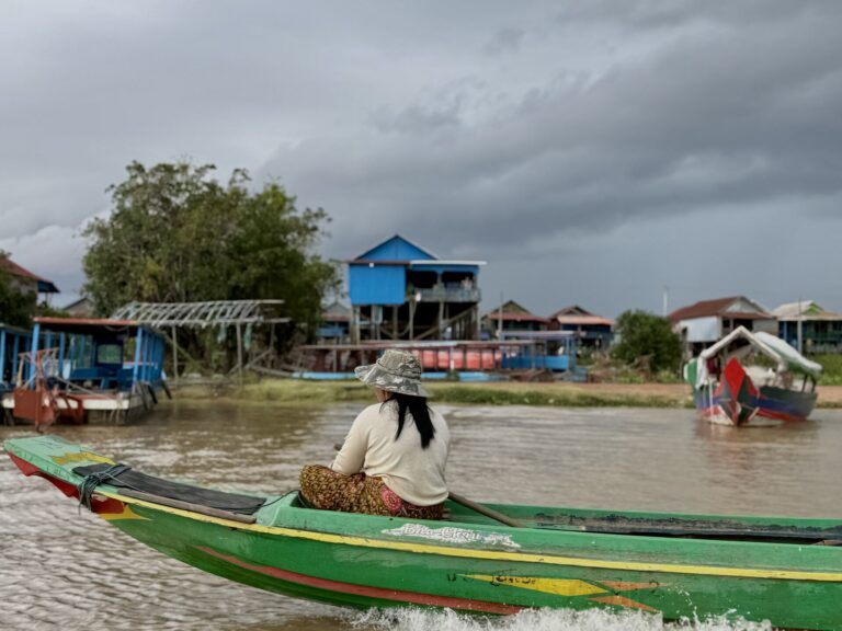 A woman rides the bow of a green boat toward Kampong Phluk under a storm sky