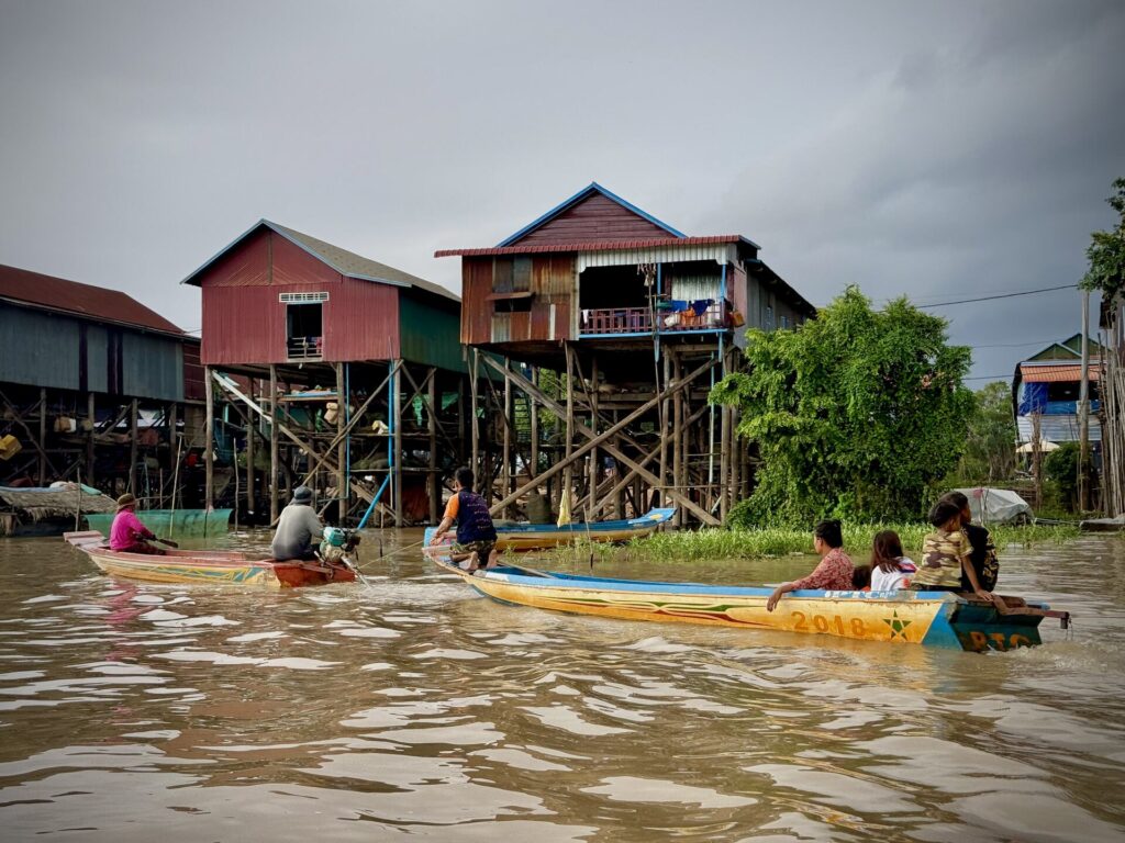 Families in wooden boats passing below the tall stilt houses of Kampong Phluk at high water