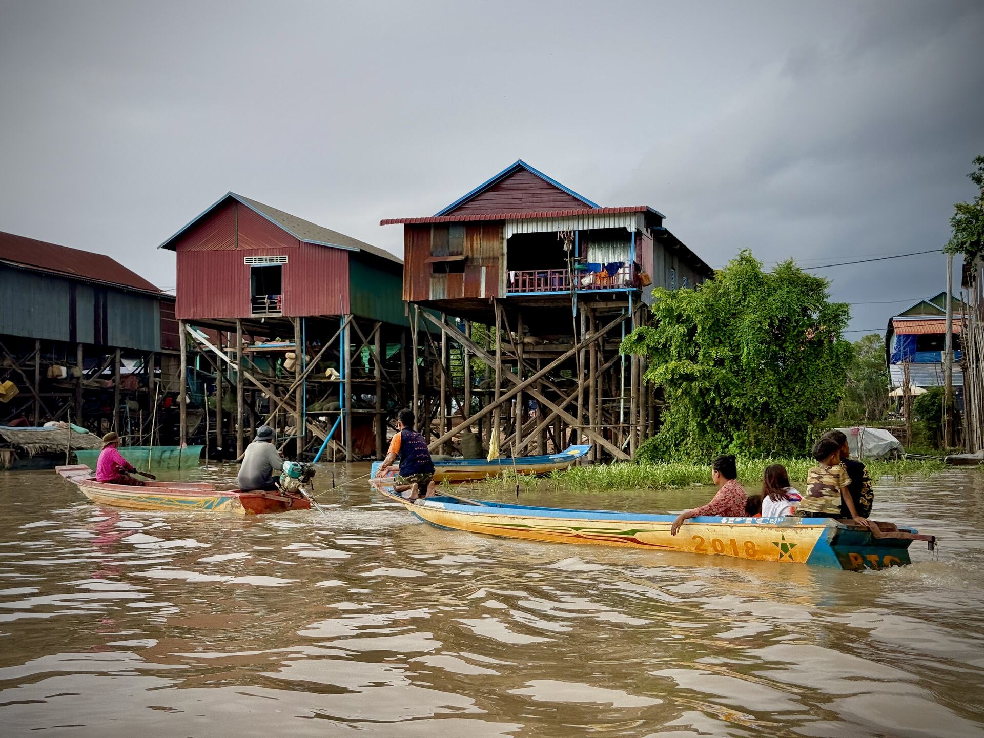 Families in wooden boats passing below the tall stilt houses of Kampong Phluk at high water