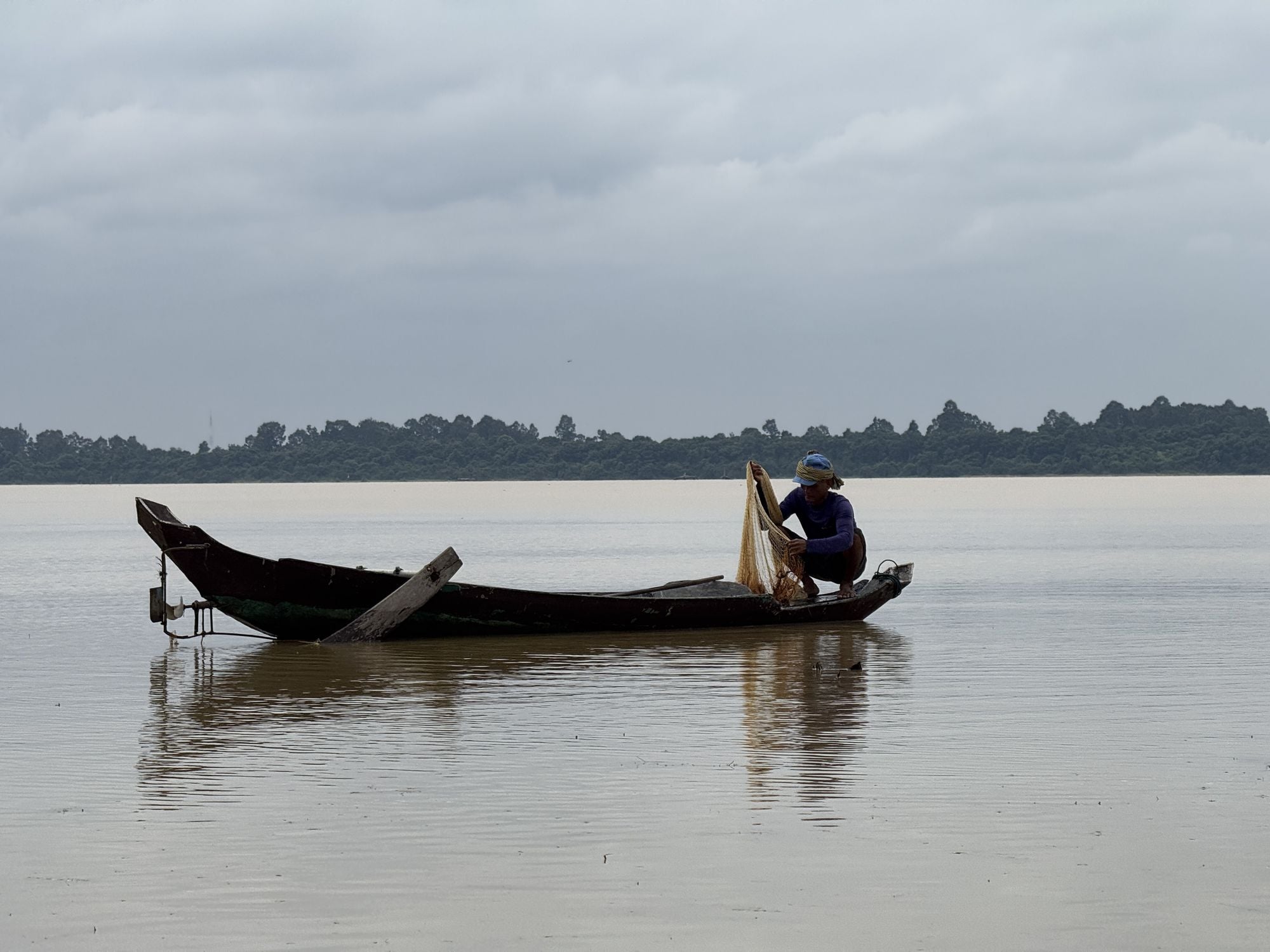 A fisherman works his net from a wooden boat on the flat water of the West Baray