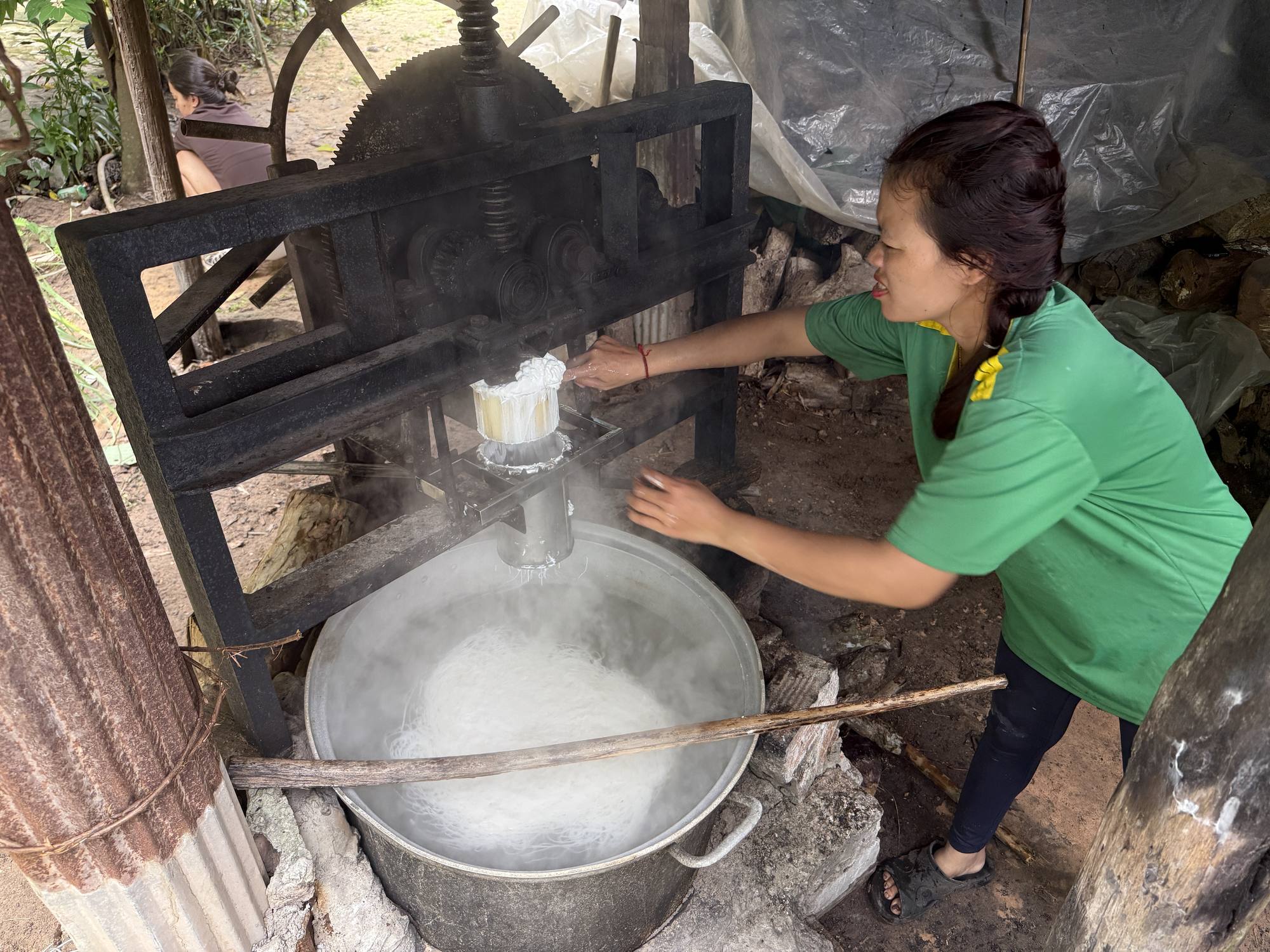 A woman works the cast-iron press that pushes rice dough into boiling water to make Num Banh Chok