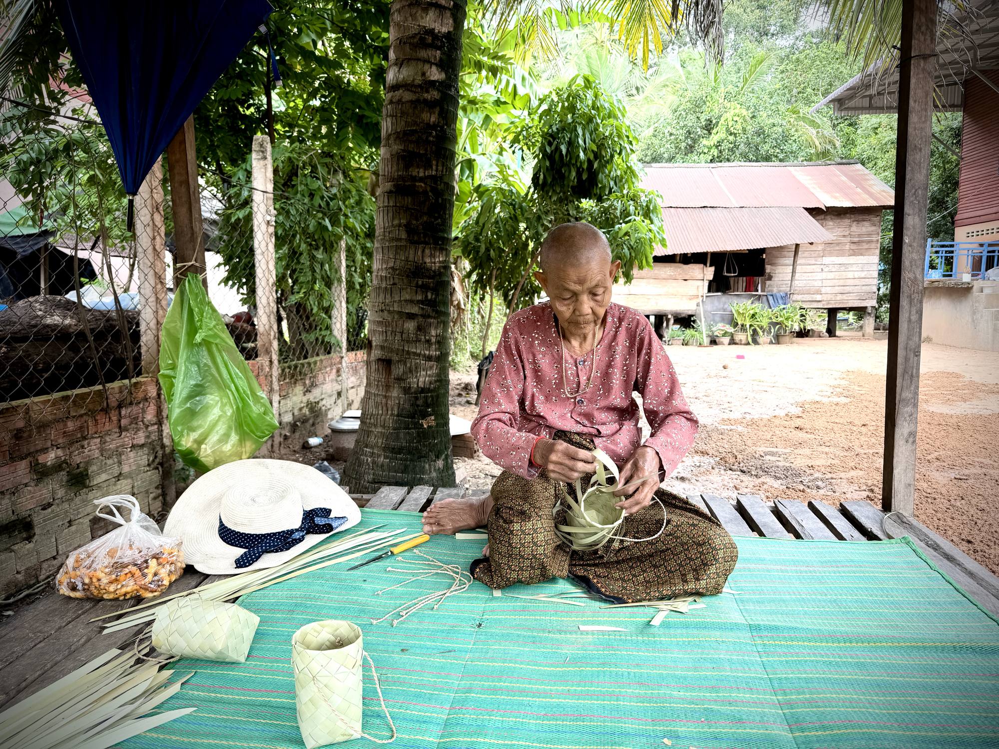 An elderly woman weaves palm leaf into smok on a mat outside her house