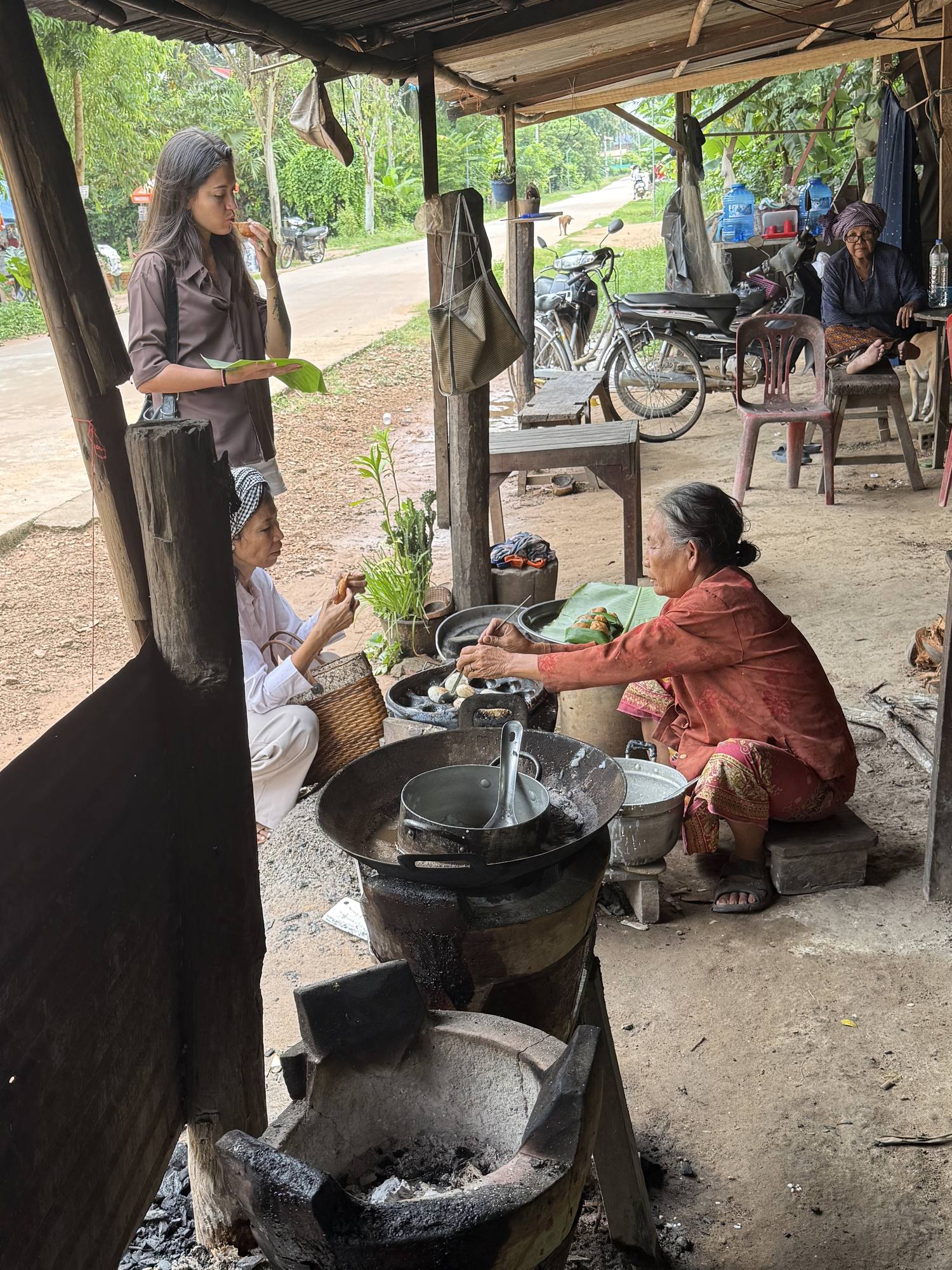 A guest stands eating at the roadside kitchen while two women cook over charcoal